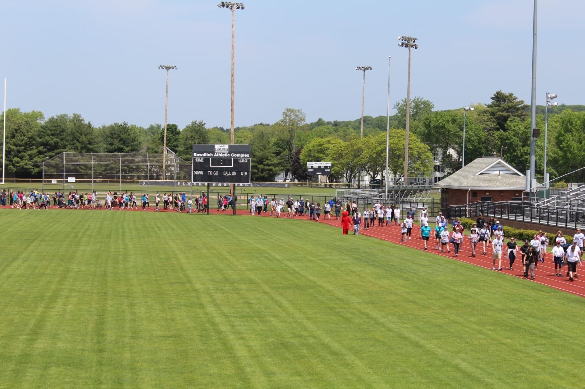 People walk around a grass field on a red track on a sunny day in May.